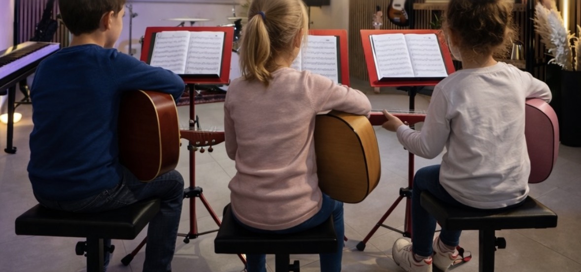 Drei Kinder sitzen von hinten gesehen mit Akustik-Gitarren vor roten Notenständern